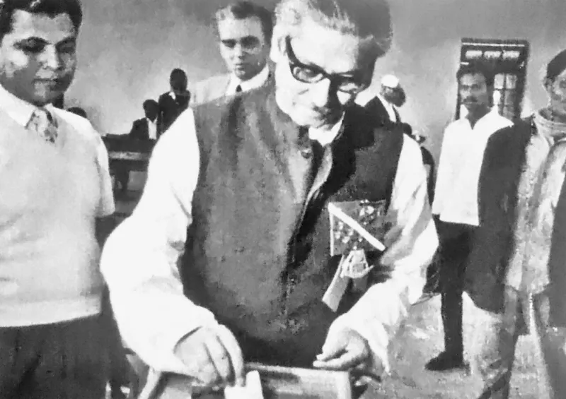 চশমা ও কালো কোট পরা শেখ মুজিবুর রহমান ব্যালট বাক্সে ভোট দিচ্ছেন, বুকে নৌকার ব্যাজ