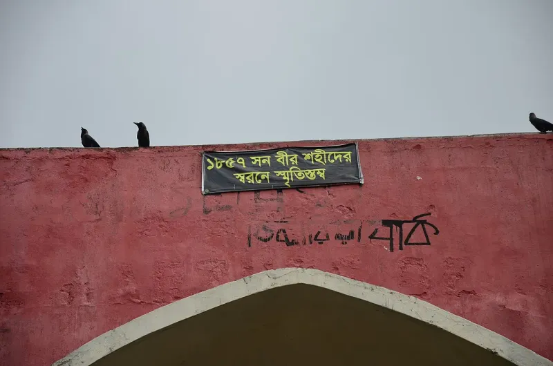 লাল দেয়ালে ‘১৮৫৭ সন বীর শহীদদের স্মরণে স্মৃতিস্তম্ভ’ লেখা ব্যানার, নিচে খিলান