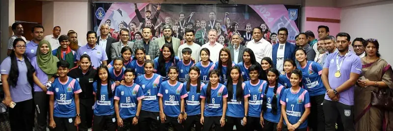 নীল জার্সি পরা নারী ফুটবলাররা কর্মকর্তাদের সঙ্গে সারি বেঁধে দাঁড়িয়ে, পেছনে শিরোপা উদ্যাপনের ব্যানার