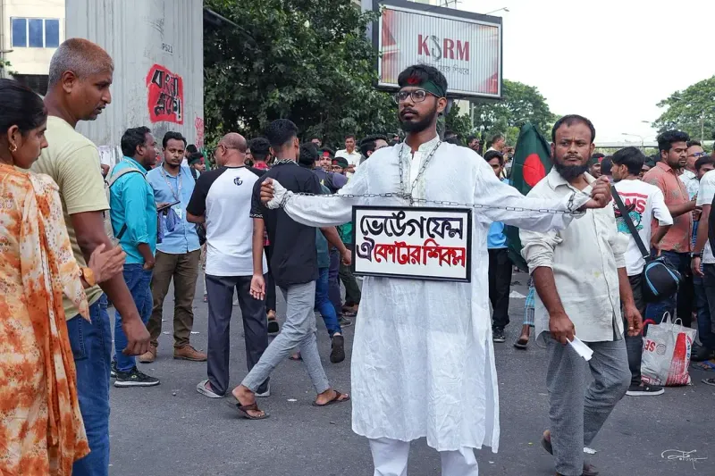 রাস্তায় শিকলে বাঁধা সাদা পাঞ্জাবি পরা এক তরুণের গলায় ‘ভেঙে ফেল কোটার শিকল’ লেখা প্ল্যাকার্ড, চারপাশে আন্দোলনকারীরা